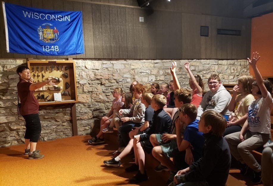 a group of fourth graders listen to a speaker at Stonefield village who is standing by some artifacts and a Wisconsin flag