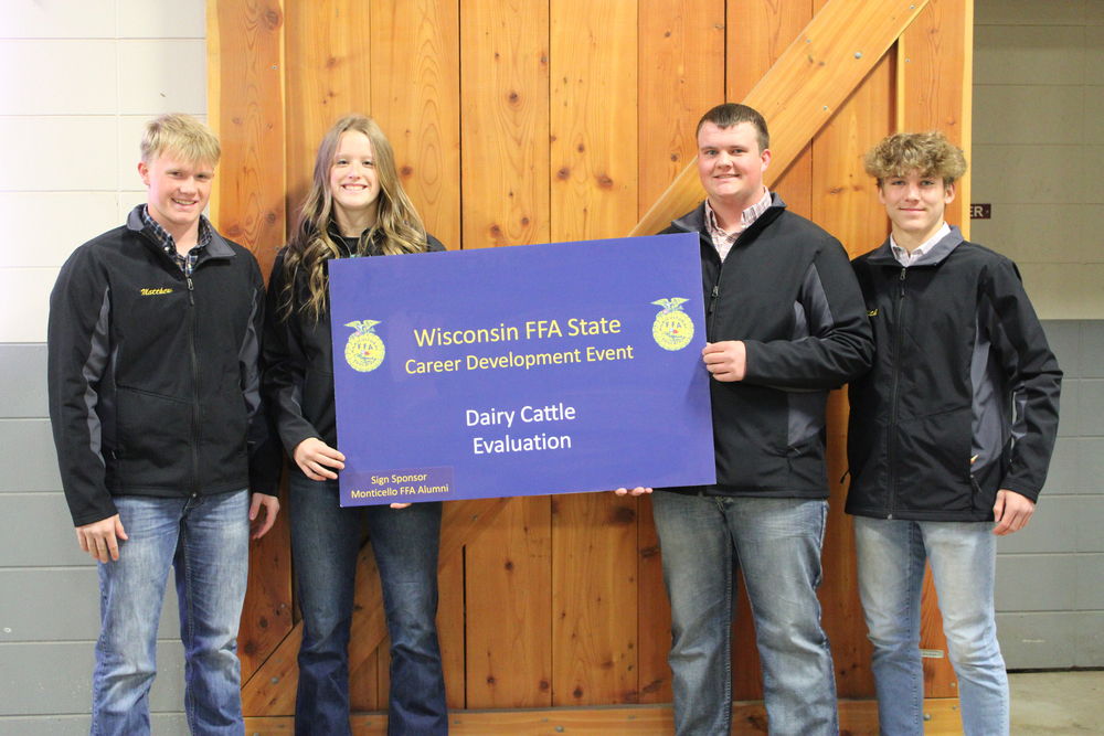 dairy team stands in front of a barn door holding blue sign