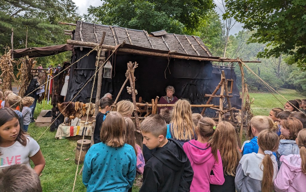 students gather in front of a longhouse and listen as someone tells them about it